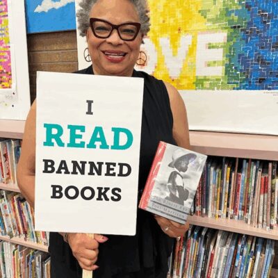 Dana Vickers Shelley is inside of a school library and holding a sign that says, "I read banned books." She is holding a copy of The Bluest Eye by Toni Morrison. Dana has short gray and black hair, is wearing cat-eye glasses and earings, and is smiling.