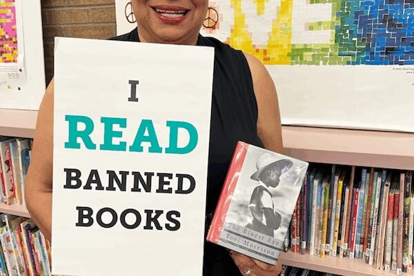 Dana Vickers Shelley is inside of a school library and holding a sign that says, "I read banned books." She is holding a copy of The Bluest Eye by Toni Morrison. Dana has short gray and black hair, is wearing cat-eye glasses and earings, and is smiling.