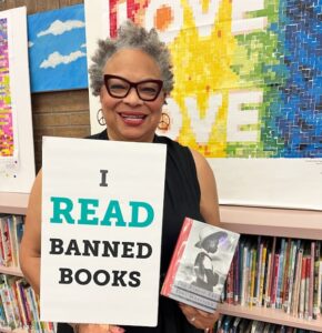 Dana Vickers Shelley is inside of a school library and holding a sign that says, "I read banned books." She is holding a copy of The Bluest Eye by Toni Morrison. Dana has short gray and black hair, is wearing cat-eye glasses and earings, and is smiling.