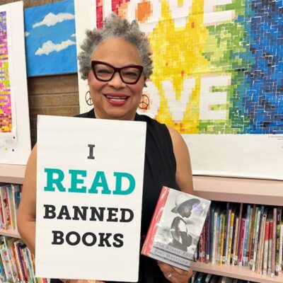 Dana Vickers Shelley is inside of a school library and holding a sign that says, "I read banned books." She is holding a copy of The Bluest Eye by Toni Morrison. Dana has short gray and black hair, is wearing cat-eye glasses and earings, and is smiling.