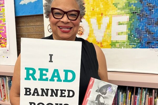 Dana Vickers Shelley is inside of a school library and holding a sign that says, "I read banned books." She is holding a copy of The Bluest Eye by Toni Morrison. Dana has short gray and black hair, is wearing cat-eye glasses and earings, and is smiling.