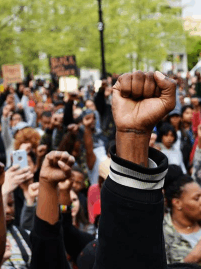 A Black person's fights in the air with a crowd of Black people behind them at a Baltimore Uprising protest