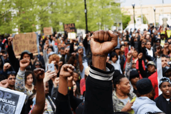 A Black person's fights in the air with a crowd of Black people behind them at a Baltimore Uprising protest