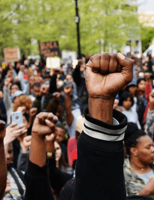 A Black person's fights in the air with a crowd of Black people behind them at a Baltimore Uprising protest