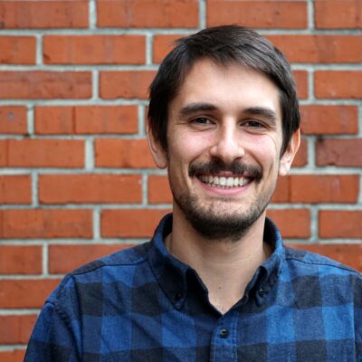 Sebastian Brown is wearing a blue and black flannel, is smiling, and is shown from the shoulders up in front of a brick wall.