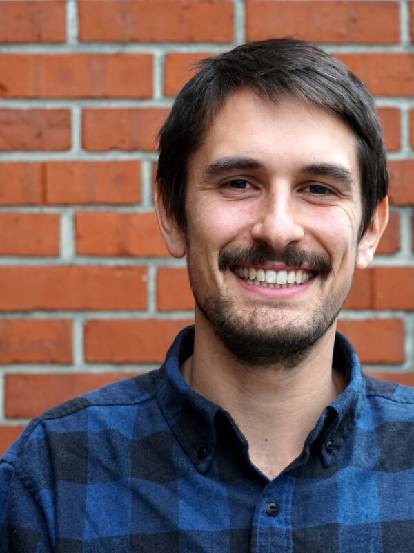 Sebastian Brown is wearing a blue and black flannel, is smiling, and is shown from the shoulders up in front of a brick wall.