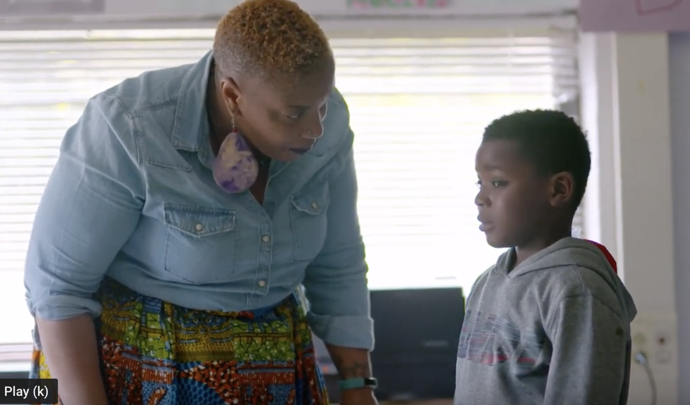 Shantay McKinily is pictured standing with and comforting a child. Shantay is wearing a blue long-sleeved shirt and colorful skirt. The student is wearing a hoodie.