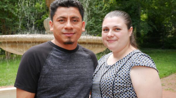 Elmer (left) and Alyse (right) Sanchez are standing in front of a water fountain with green trees behind them.