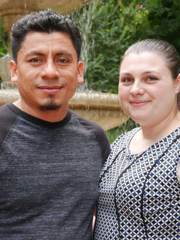 Elmer (left) and Alyse (right) Sanchez are standing in front of a water fountain with green trees behind them.