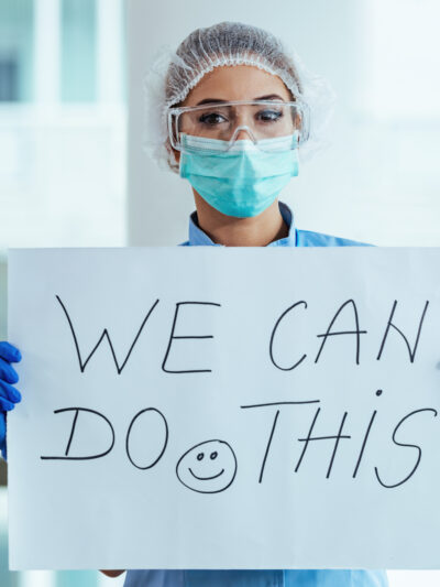 Healthcare worker that has goggles, a face mask, and gloves is holding a sign that says, "We can do this" with a smiley face.