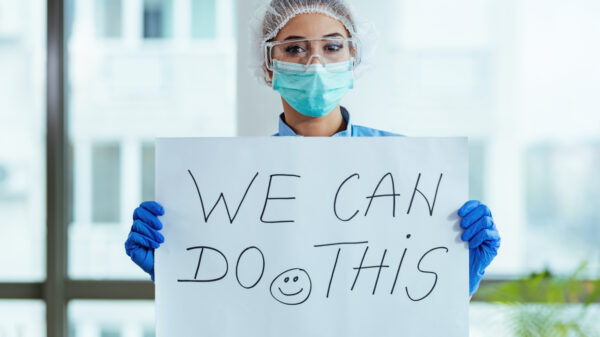 Healthcare worker that has goggles, a face mask, and gloves is holding a sign that says, "We can do this" with a smiley face.