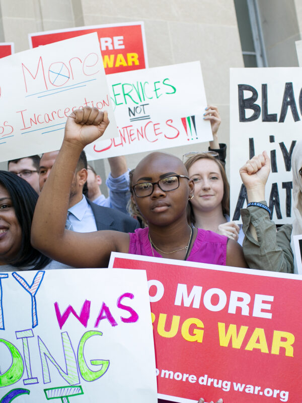 Protest with a Black person in the center with their fist raised. The signs say "no more drug war" and "Black Lives Matter" and "resist".