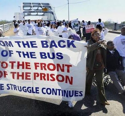 Black marchers on the Edmund Pettus Bridge holding a banner that says "From the back of the bus ot the front of the prison, the struggle continues."