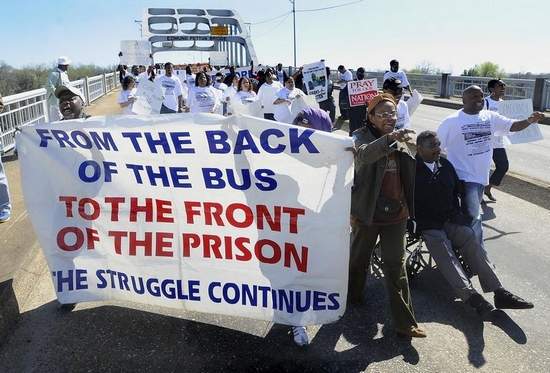Black marchers on the Edmund Pettus Bridge holding a banner that says "From the back of the bus ot the front of the prison, the struggle continues."