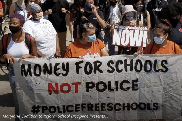The picture is of people at a protest march holding a big banner that says "Money For Schools Not Police. Police Free Schools".