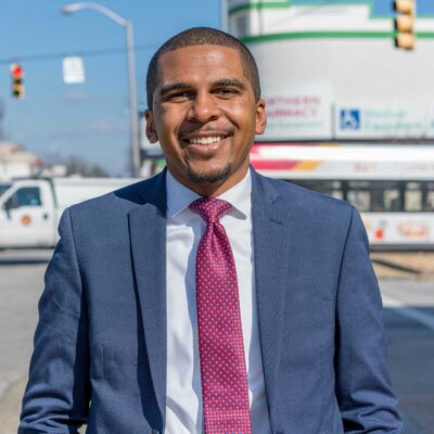 Image shows a picture of Caylin Young in a blue suit and fuschia time. He is standing on a Baltimore City street on a sunny day. He is a Black may with short hair and he is smiling in the photo.