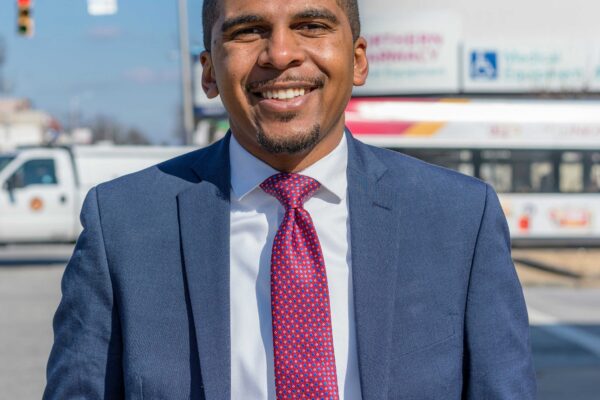 Image shows a picture of Caylin Young in a blue suit and fuschia time. He is standing on a Baltimore City street on a sunny day. He is a Black may with short hair and he is smiling in the photo.