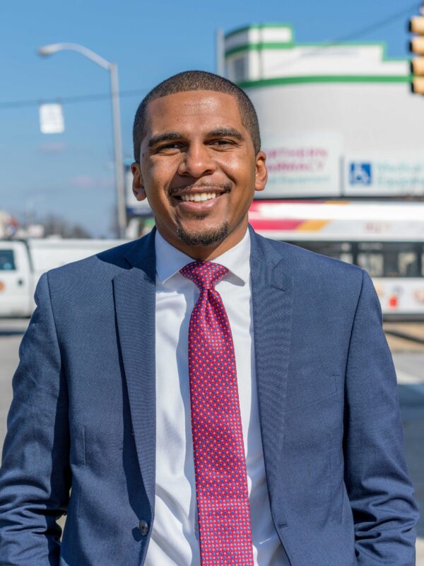 Image shows a picture of Caylin Young in a blue suit and fuschia time. He is standing on a Baltimore City street on a sunny day. He is a Black may with short hair and he is smiling in the photo.