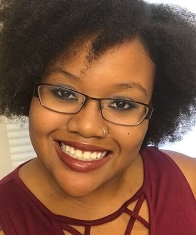 Picture shows Kristen Lundy, a Black Latina, wearing glasses and a plum sleeveless shirt. She is smiling.