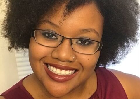 Picture shows Kristen Lundy, a Black Latina, wearing glasses and a plum sleeveless shirt. She is smiling.