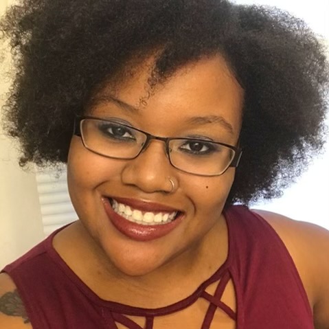 Picture shows Kristen Lundy, a Black Latina, wearing glasses and a plum sleeveless shirt. She is smiling.