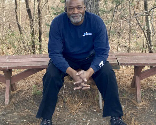 Picture of Calvin McNeill, a Black man with bald head, sitting on a bench in front of a tree.