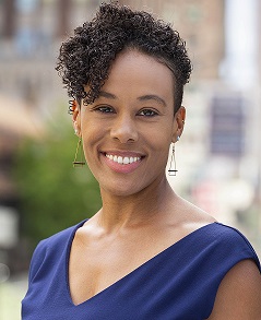 Adria Crutchfield is a Black woman with short curly hair in this picture. She is wearing a navy v-neck blue shirt and is standing outside in front of a tree.