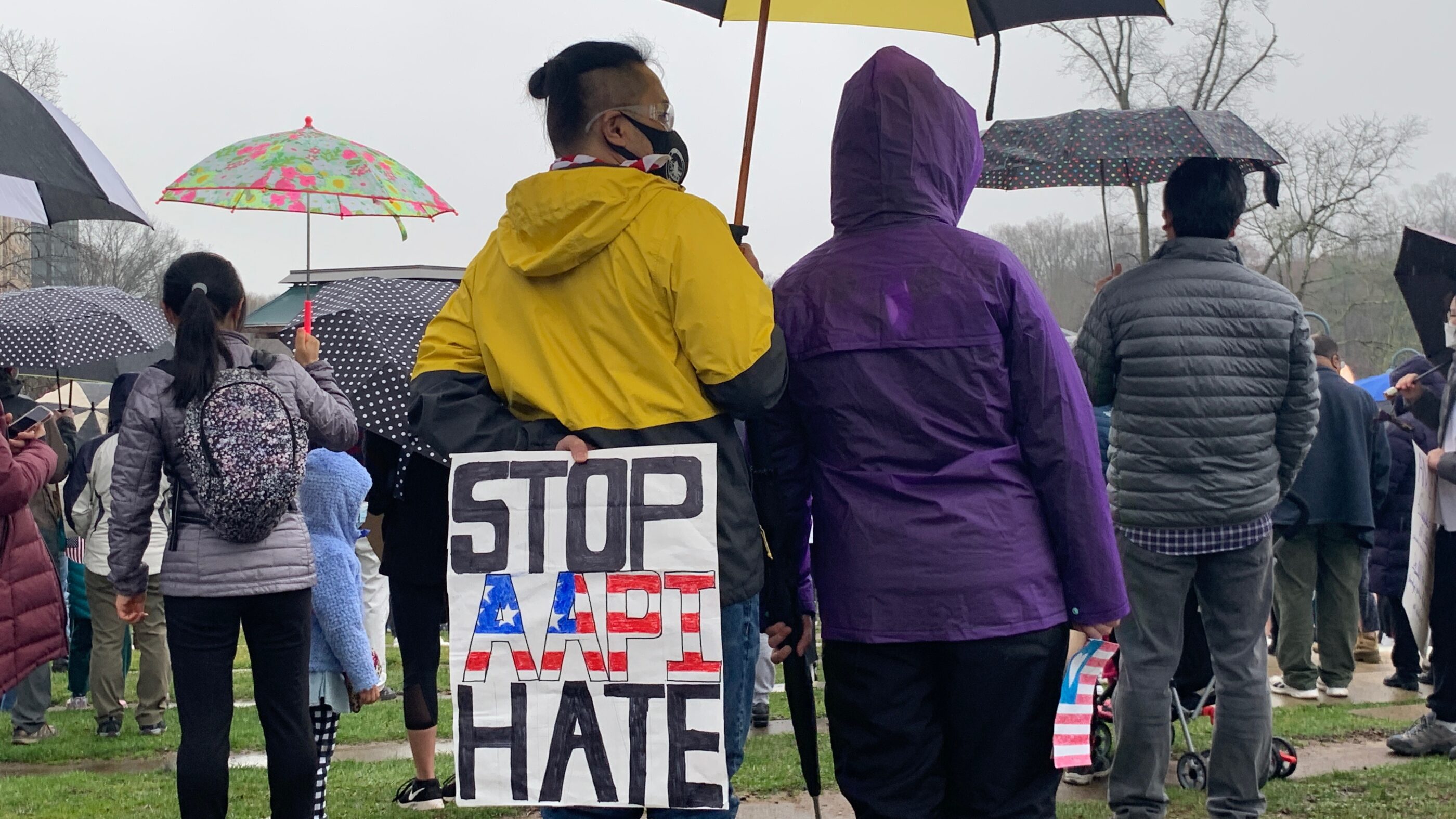 A person is standing at a rally in Columbia, Maryland, in a yellow hooded jacket, holding a yellow and black umbrella. The person is holidng a sign behind their back that says "Stop AAPI Hate". There are many other people in the picture with umbrellas.