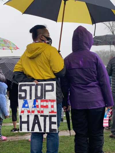 A person is standing at a rally in Columbia, Maryland, in a yellow hooded jacket, holding a yellow and black umbrella. The person is holidng a sign behind their back that says "Stop AAPI Hate". There are many other people in the picture with umbrellas.