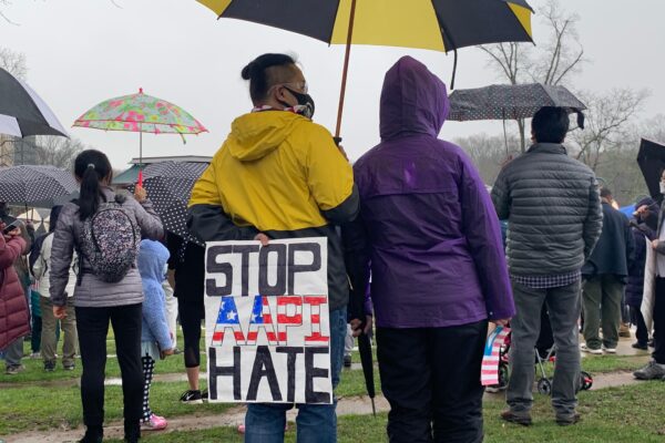 A person is standing at a rally in Columbia, Maryland, in a yellow hooded jacket, holding a yellow and black umbrella. The person is holidng a sign behind their back that says "Stop AAPI Hate". There are many other people in the picture with umbrellas.