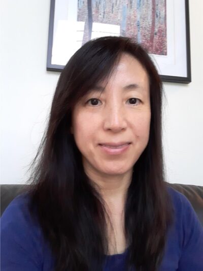 Ying Matties is a Chinese woman with long dark hair. She is wearing a dark blue top and is sitting down. There is a framed picture on the wall behind her.