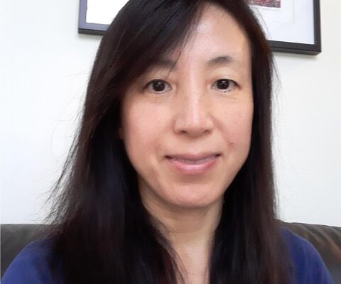 Ying Matties is a Chinese woman with long dark hair. She is wearing a dark blue top and is sitting down. There is a framed picture on the wall behind her.