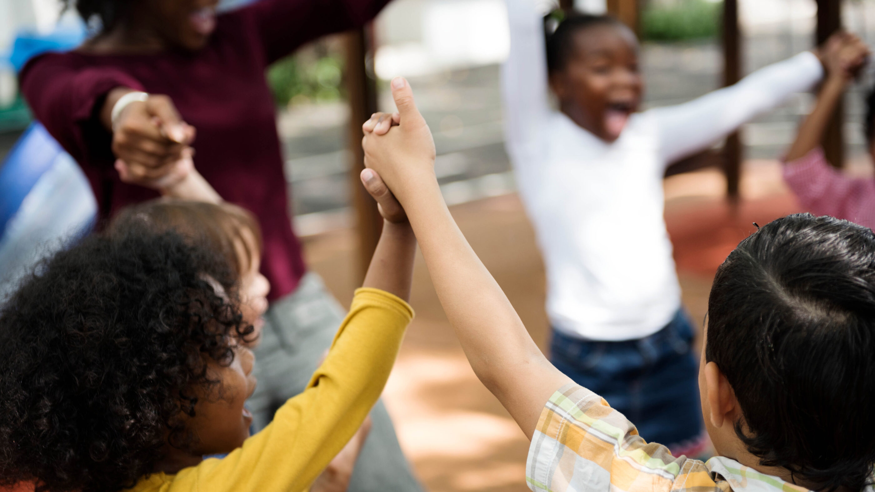 Several children are in a circle holding lifted up hands and are with an adult. They are all smiling and laughing.