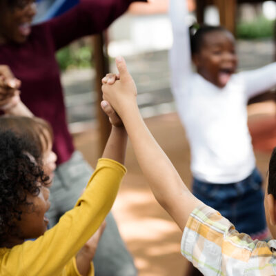 Several children are in a circle holding lifted up hands and are with an adult. They are all smiling and laughing.