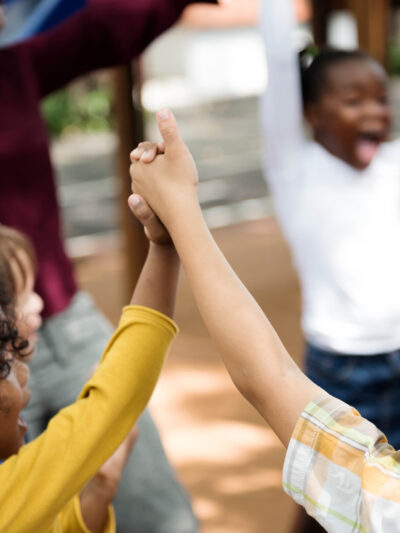 Several children are in a circle holding lifted up hands and are with an adult. They are all smiling and laughing.