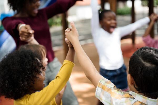 Several children are in a circle holding lifted up hands and are with an adult. They are all smiling and laughing.