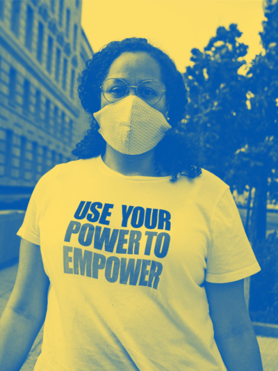 A Black person is standing on a sidewalk and has hair past their shoulders and wearing a mask and a t-shirt that says "Use Your Power to Empower."