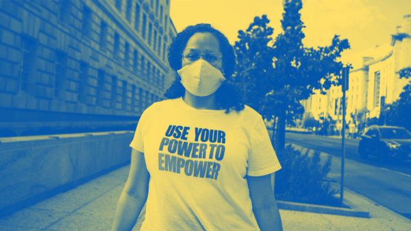 A Black person is standing on a sidewalk and has hair past their shoulders and wearing a mask and a t-shirt that says "Use Your Power to Empower."