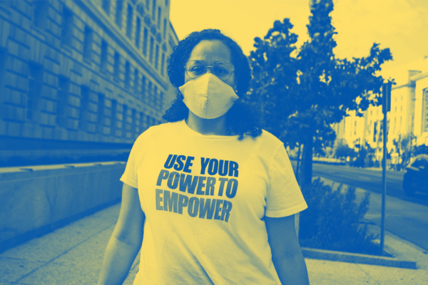 A Black person is standing on a sidewalk and has hair past their shoulders and wearing a mask and a t-shirt that says "Use Your Power to Empower."