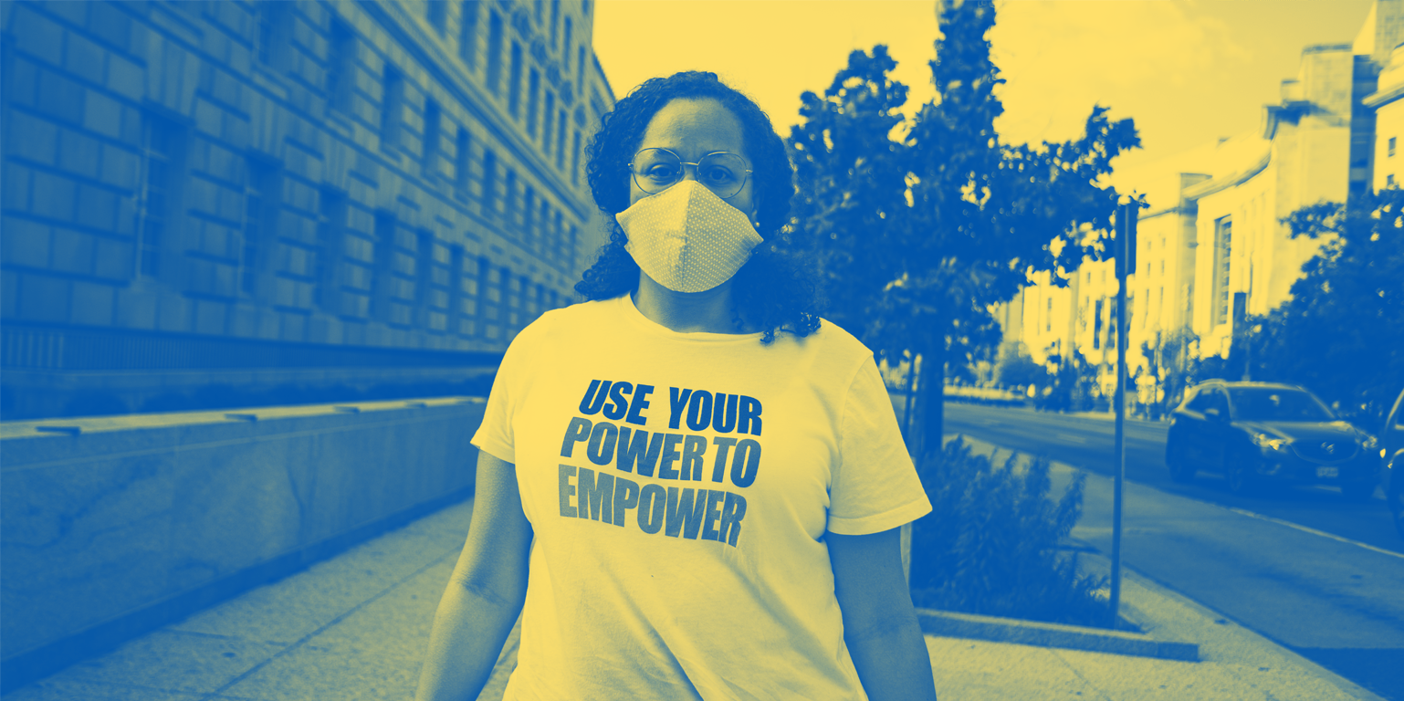 A Black person is standing on a sidewalk and has hair past their shoulders and wearing a mask and a t-shirt that says "Use Your Power to Empower."