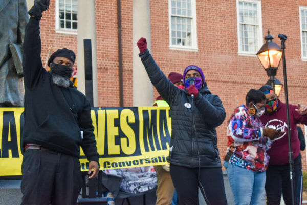 Two Black members of CJSJ have raised fists and stand in front of a Black Lives Matter banner in Annapolis at a police reform rally.