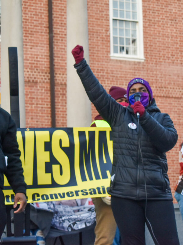 Two Black members of CJSJ have raised fists and stand in front of a Black Lives Matter banner in Annapolis at a police reform rally.
