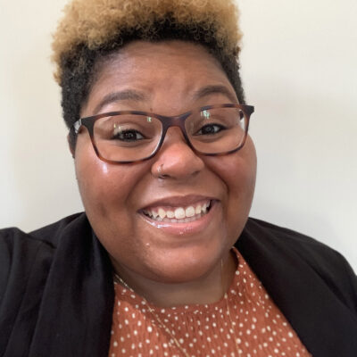 Alica Smith is a Black person, pictured with short black and bleached hair, wearing glasses, smiling, and a dark blazer over a brownish-orange shirt.