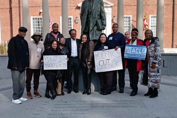 Group of advocates celebrate the veto override of parole reform in Annapolis, Maryland. They stand in front of the Thurgood Marshall statue. Some hold signs about getting politics out of parole and one person holds a sign with the ACLU-MD logo.