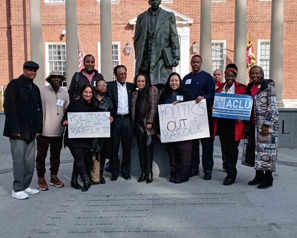 Group of advocates celebrate the veto override of parole reform in Annapolis, Maryland. They stand in front of the Thurgood Marshall statue. Some hold signs about getting politics out of parole and one person holds a sign with the ACLU-MD logo.