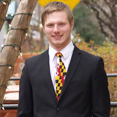 Jared is a white man with lighg colored short hair, standing in front of a tree with lights on the branches, and he is wearing a dark suit, white shirt, and Maryland flag tie. He is smiling and looking at the camera.