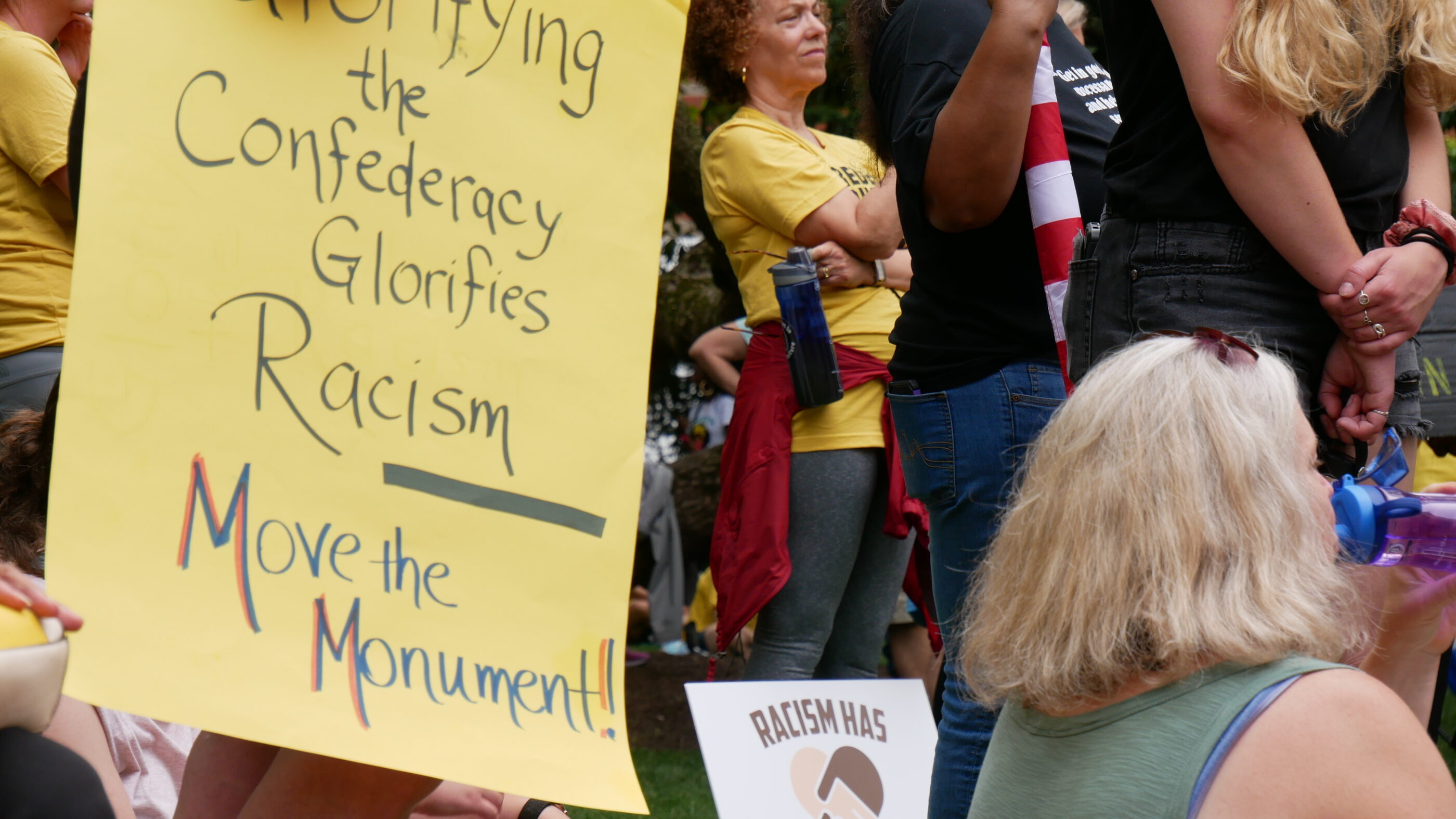 Person holds a sign that says, "Glorifying the Confederacy Glorifies Racism. Move the Monument." Another sign is visible that says, "Racism has no business here."