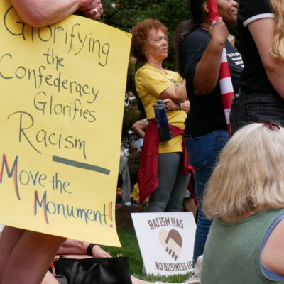 Person holds a sign that says, "Glorifying the Confederacy Glorifies Racism. Move the Monument." Another sign is visible that says, "Racism has no business here."