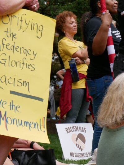 Person holds a sign that says, "Glorifying the Confederacy Glorifies Racism. Move the Monument." Another sign is visible that says, "Racism has no business here."