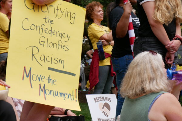 Person holds a sign that says, "Glorifying the Confederacy Glorifies Racism. Move the Monument." Another sign is visible that says, "Racism has no business here."